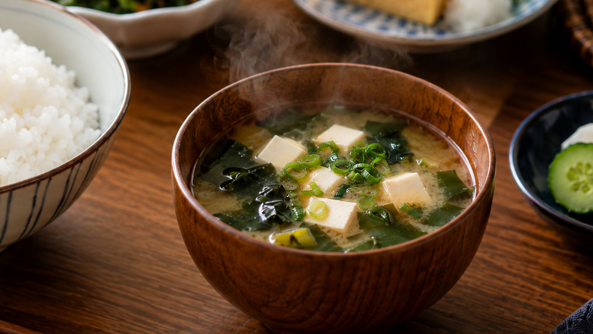 A bowl of miso soup with tofu, wakame seaweed, and green onions, served with rice and pickles as part of a Japanese home meal.
