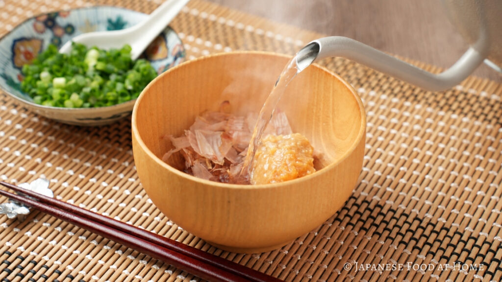 Hot water being poured over miso and bonito flakes in a wooden bowl to make kachuyu, an Okinawan miso soup.