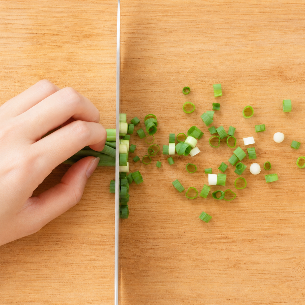 Green onion being thinly sliced crosswise into small rounds on a wooden cutting board
