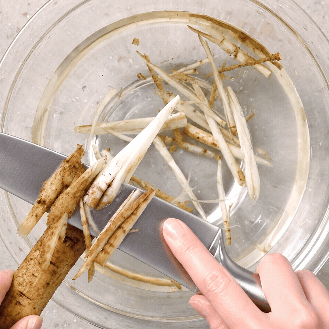 Gobo being shaved into thin pieces over a bowl of water using the sasagaki technique