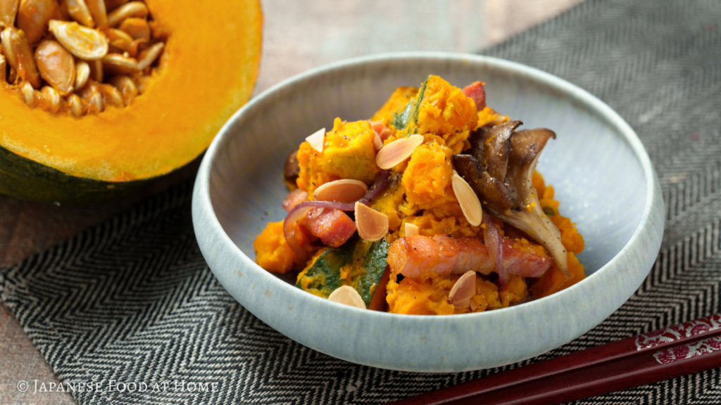Japanese-style kabocha salad with bacon, maitake mushrooms, red onion, and toasted sliced almonds, served in a ceramic bowl with fresh kabocha squash in the background.